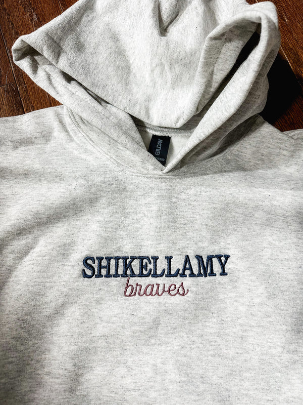 Gray hoodie with 'SHIKELLAMY braves' text on a wooden surface